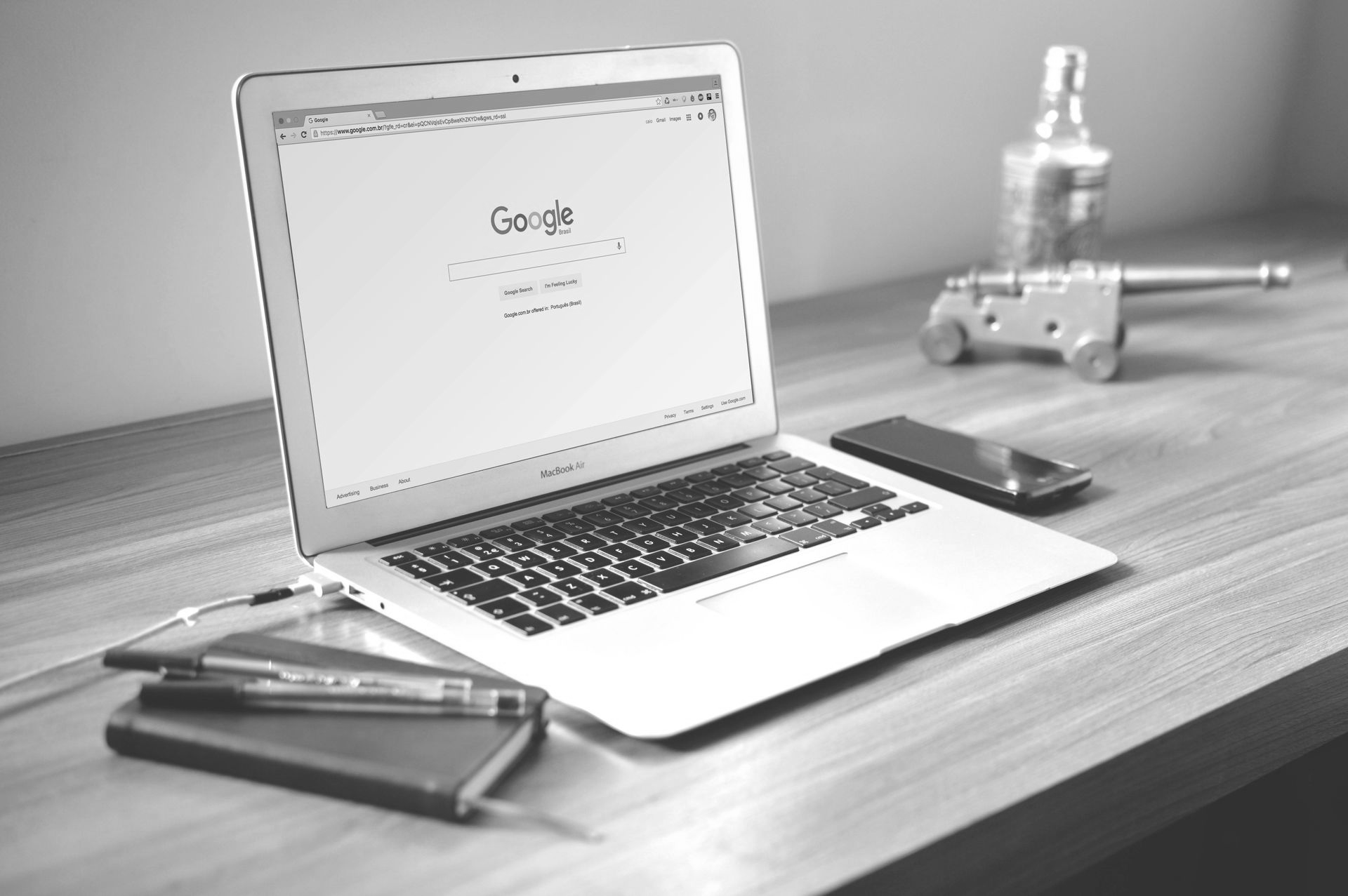A laptop open to a Google search page sits on a wooden desk with a notebook, pens, a smartphone, and a model cannon.