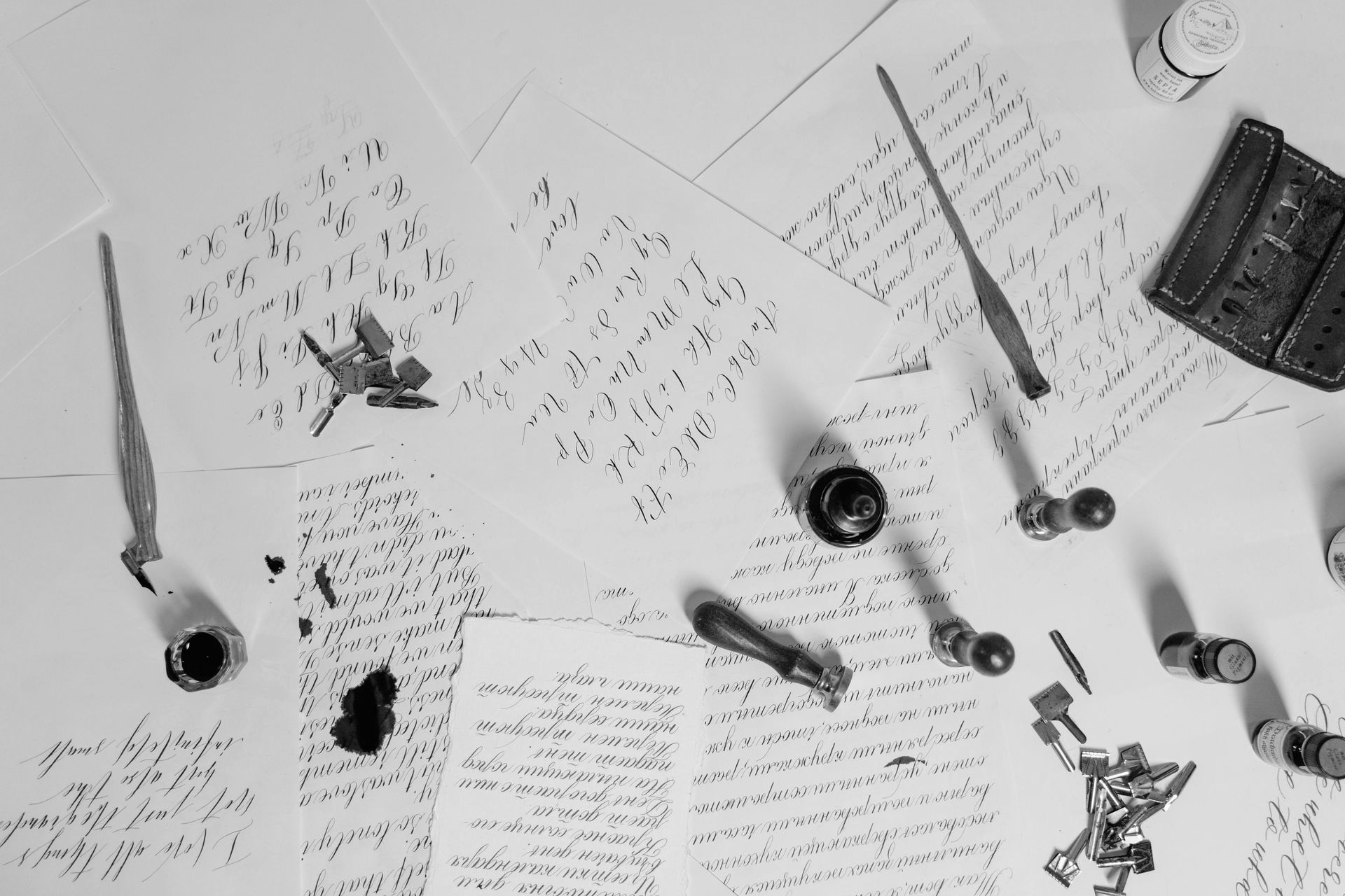 A black and white overhead view of fountain pens, ink bottles, and handwritten calligraphy on scattered paper sheets.