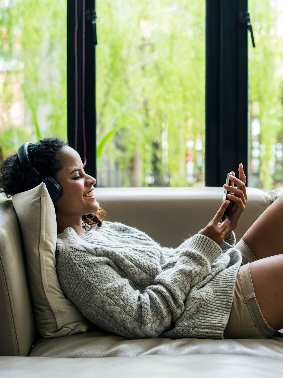 A woman is laying on a couch wearing headphones and looking at her phone.