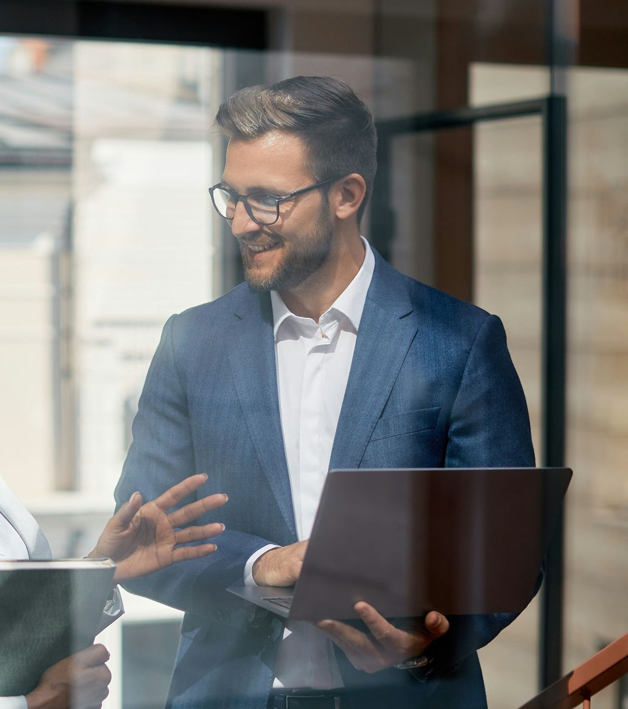 A man in a suit is holding a laptop and talking to another man.