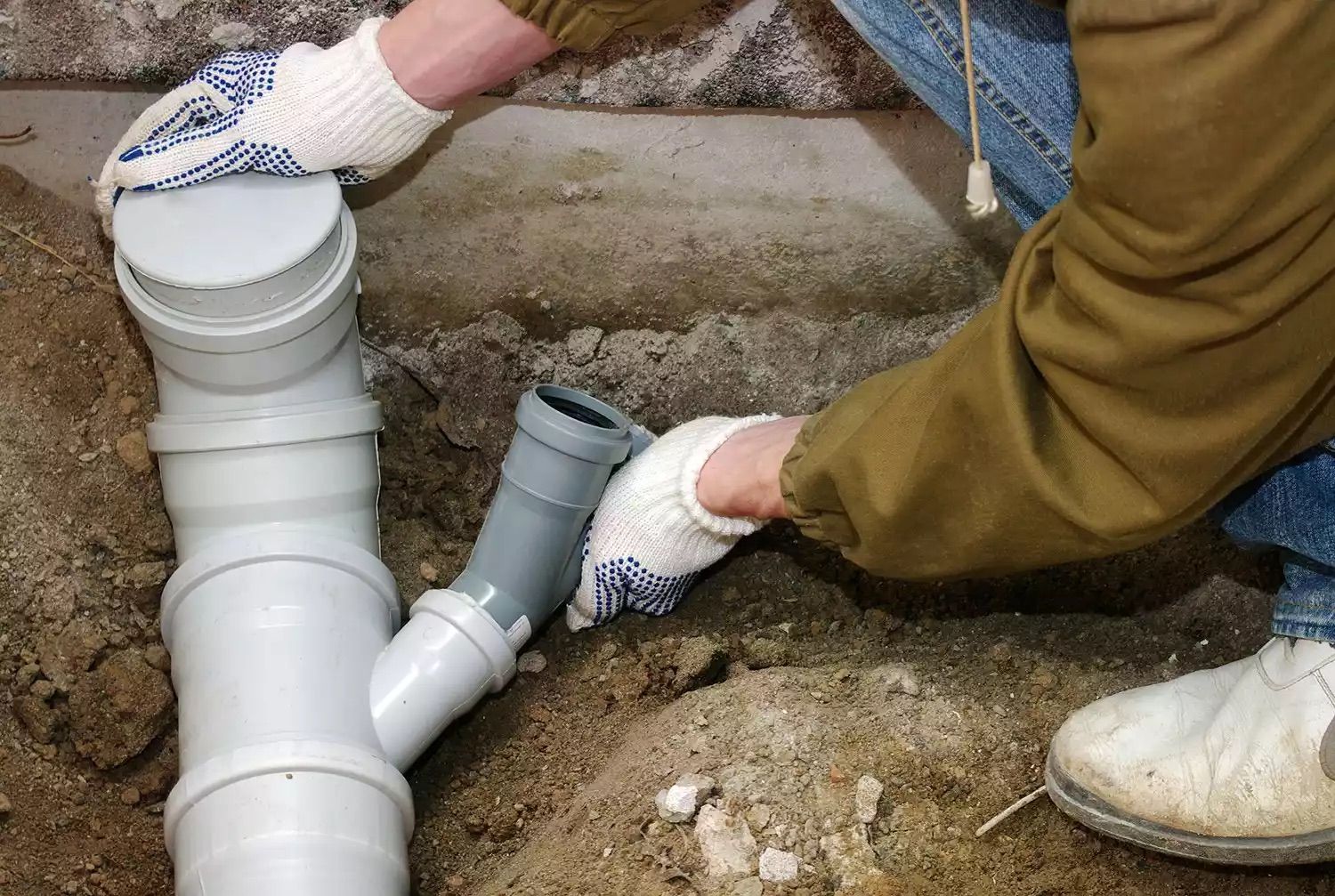 Person connecting gray pipe to white plumbing in a trench. Hands wear gloves. Outdoor pipe repair.