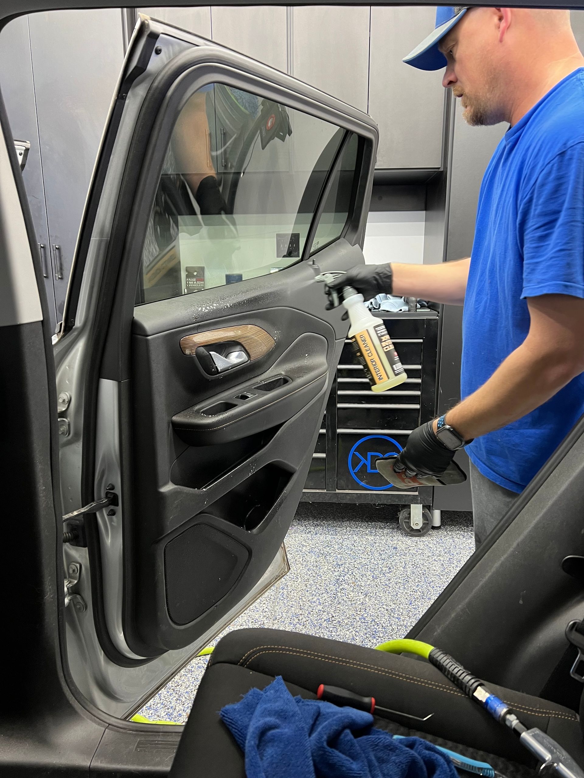 Person sprays cleaning solution on a car door panel, detailing interior. Black gloves, blue shirt.