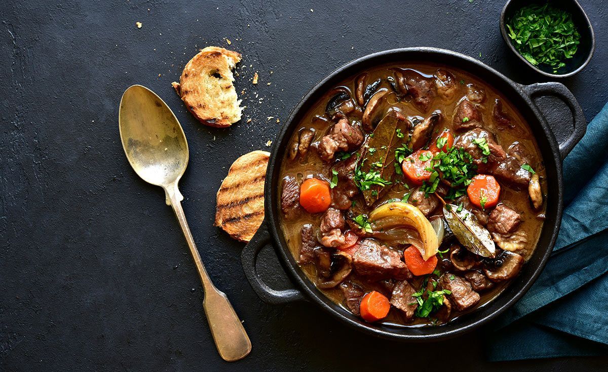 A bowl of beef stew with carrots and mushrooms next to a spoon and a piece of bread.