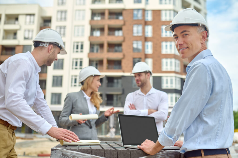 A group of construction workers are standing around a laptop on a construction site.