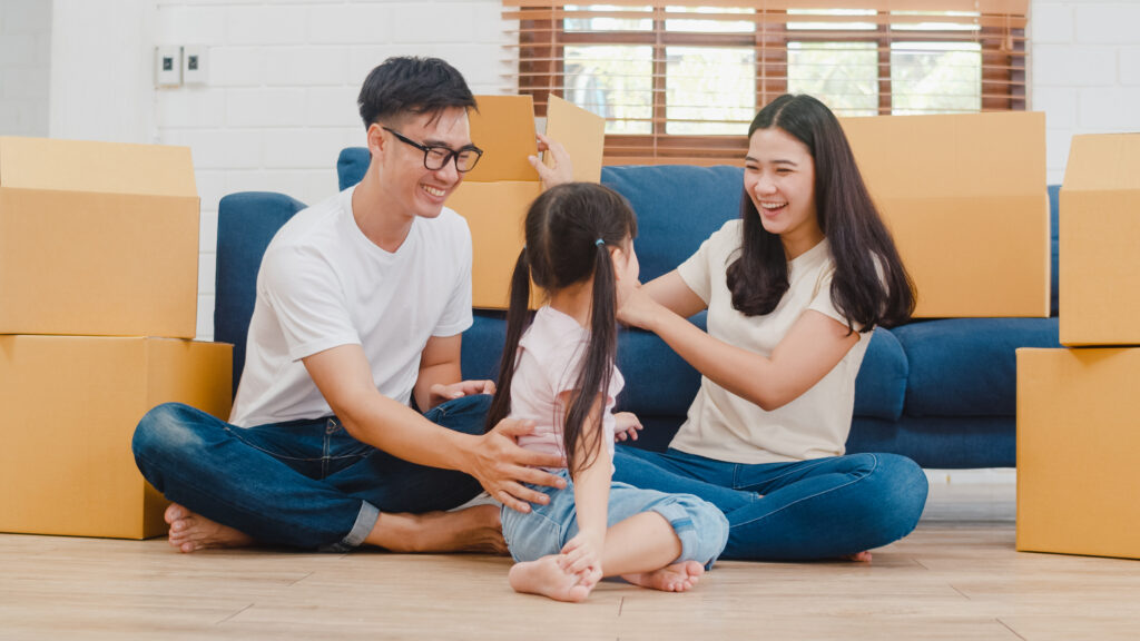 A family is sitting on the floor in a living room surrounded by cardboard boxes.