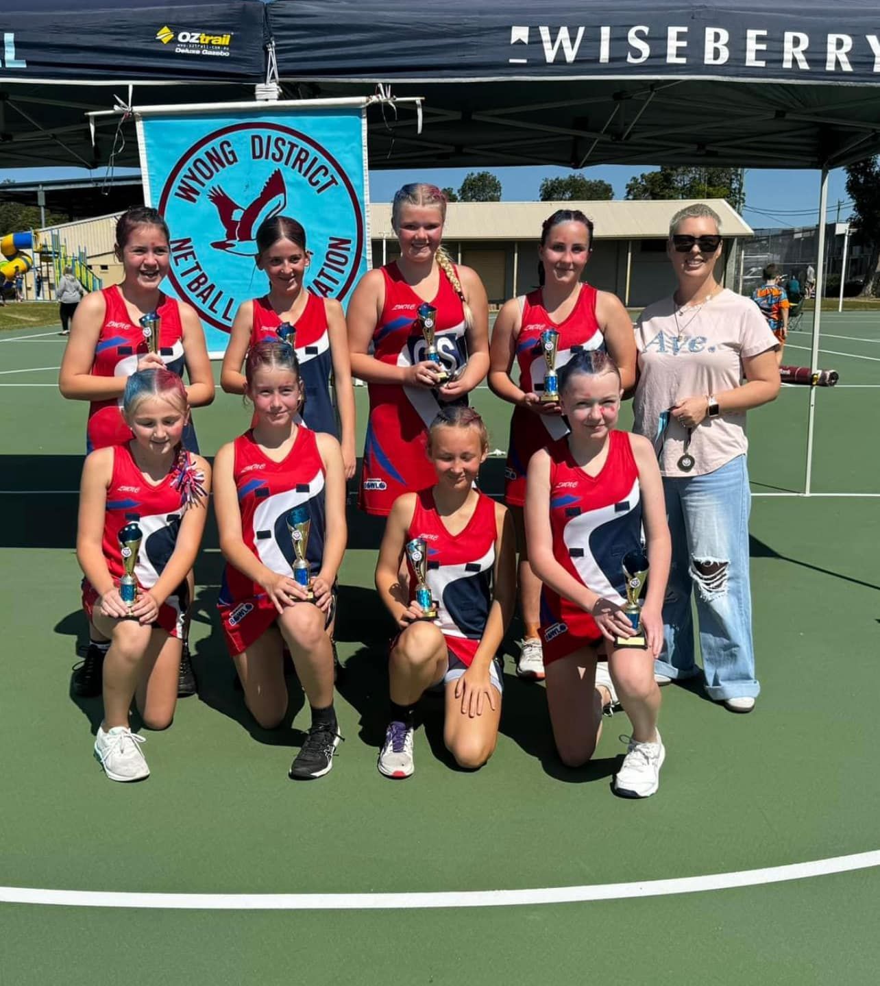 A netball team wearing red, white, and blue uniforms poses with trophies on a court under a banner.