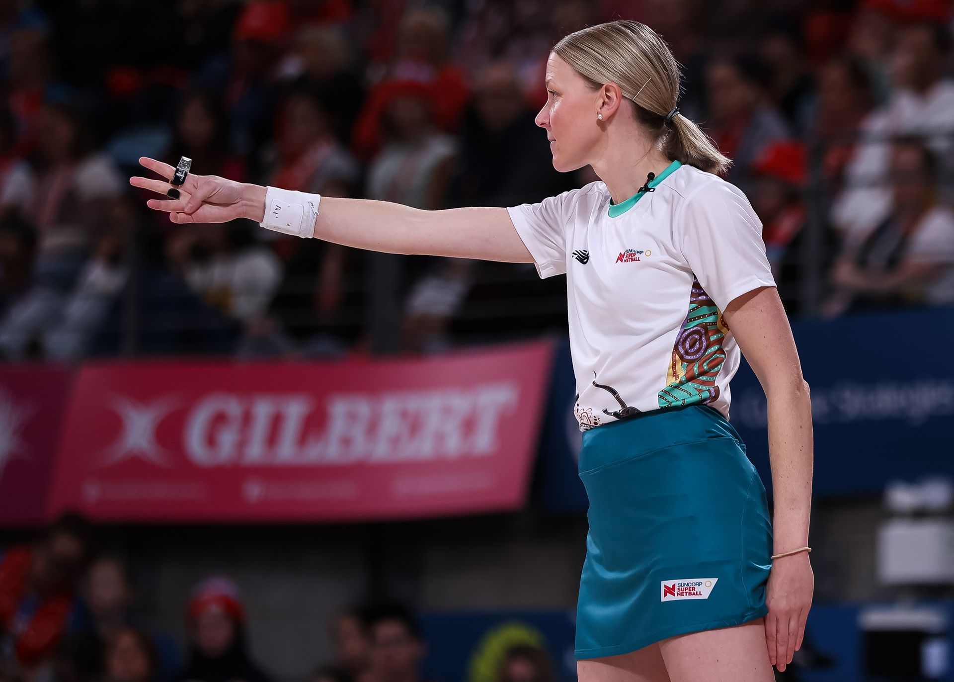 A netball umpire in a white and teal uniform stands on a court, signaling with her arm outstretched.
