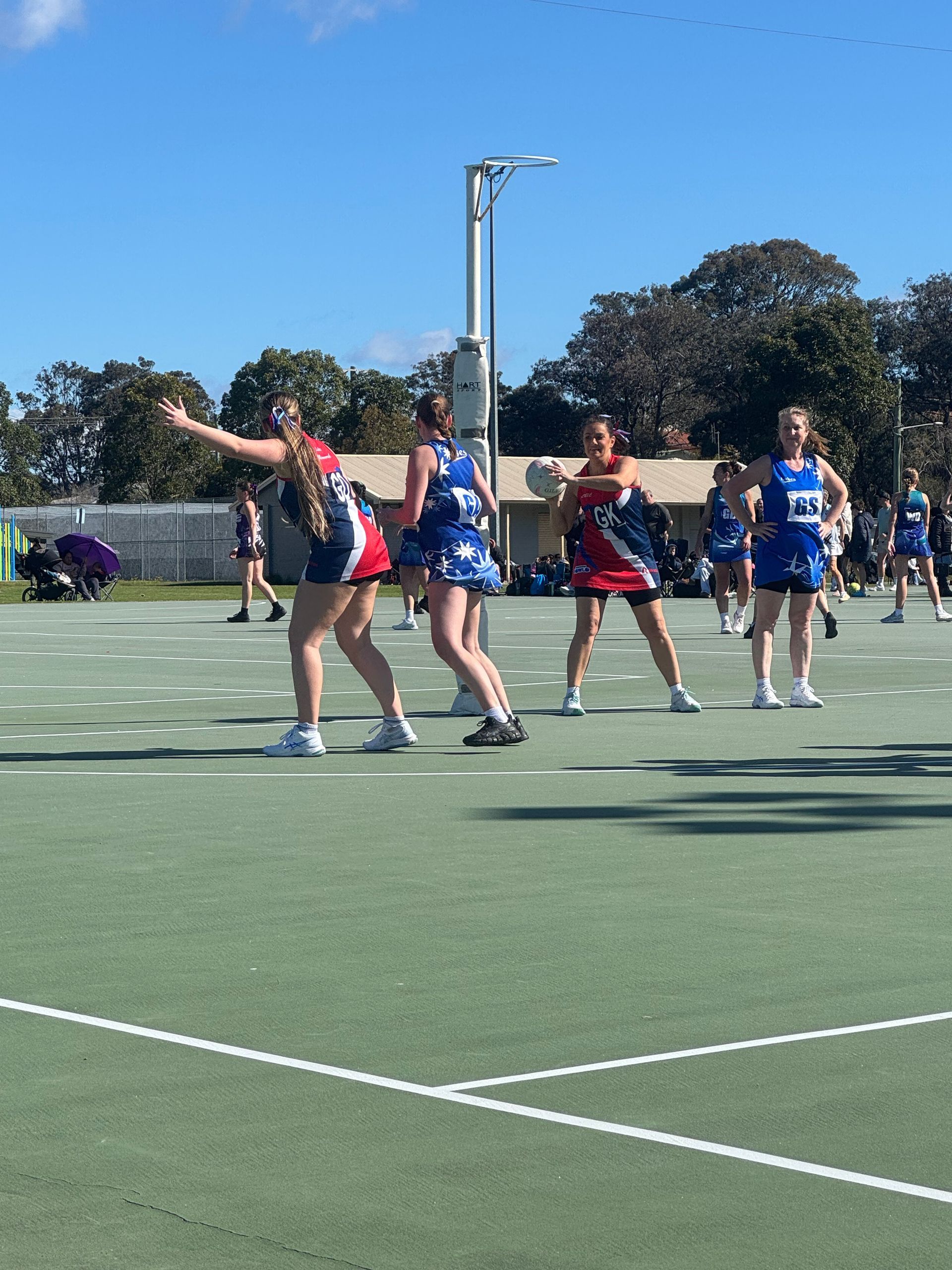 Netball players in red, white, and blue uniforms playing on an outdoor court under a clear blue sky.
