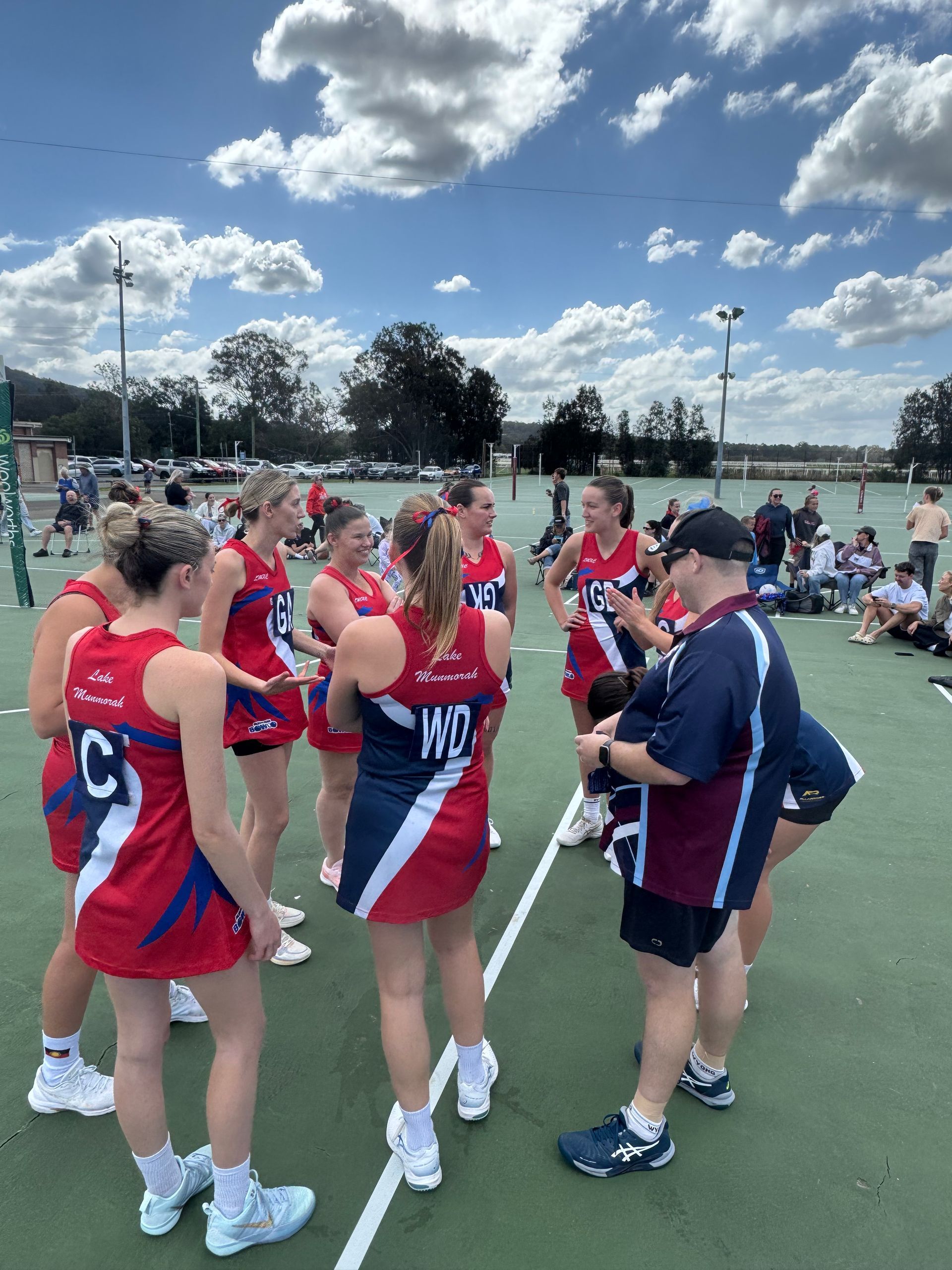 A sports team in red, white, and navy uniforms gathers in a circle on an outdoor court to listen to a coach.