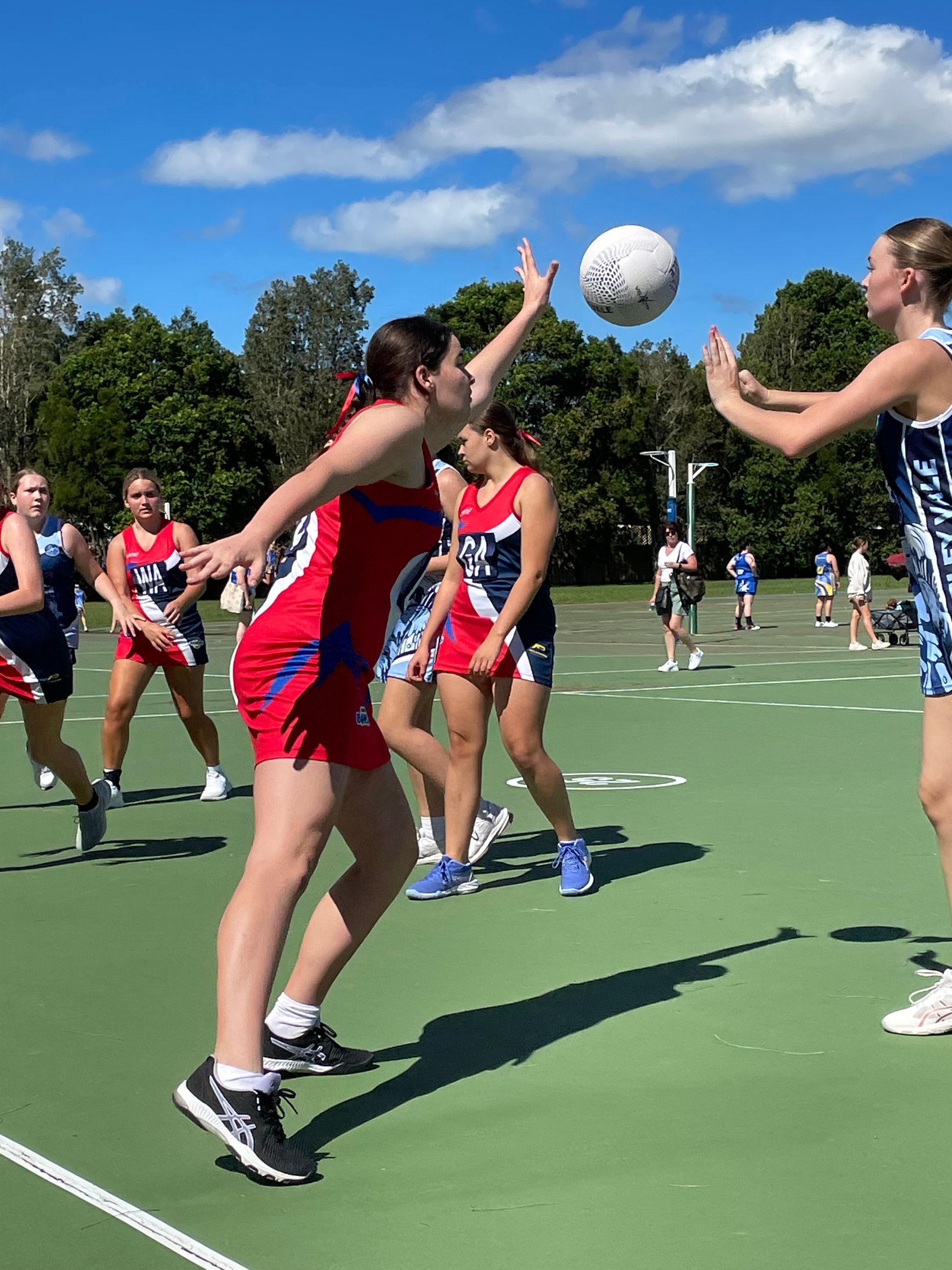 Athletes playing netball on an outdoor court, one player reaching up to catch a ball near another player.