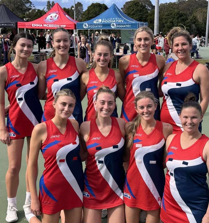 A group of nine netball players in matching red, white, and blue uniforms pose together on an outdoor court.