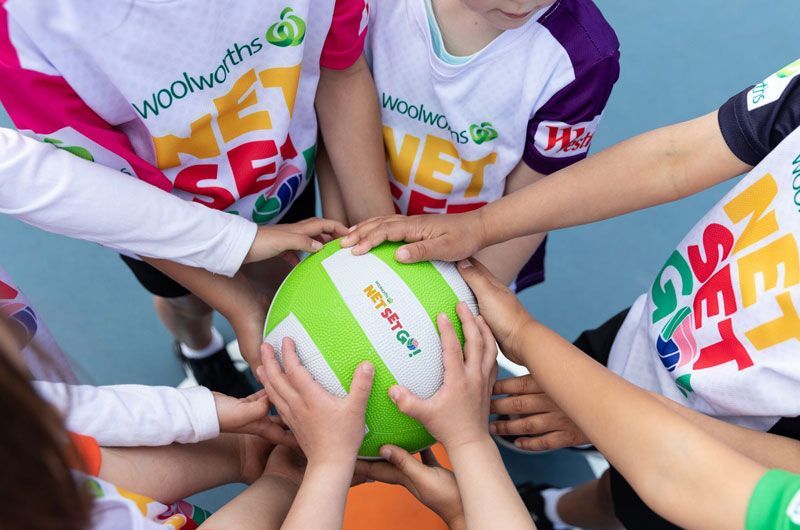 Several children wearing Woolworths NetSetGO jerseys place their hands together on a green and white netball.