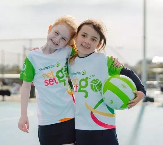 Two smiling children in matching green and white sports uniforms stand on an outdoor court, one holding a green netball.
