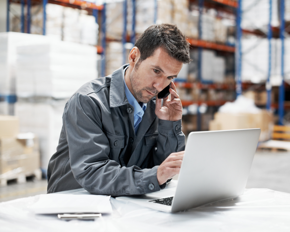 A man is talking on a cell phone while using a laptop computer in a warehouse.
