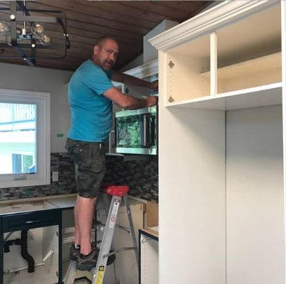 A man is standing on a ladder in a kitchen working on a microwave