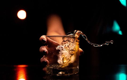 Hand holding a whiskey glass with ice and a twist of citrus in a dark bar-like setting