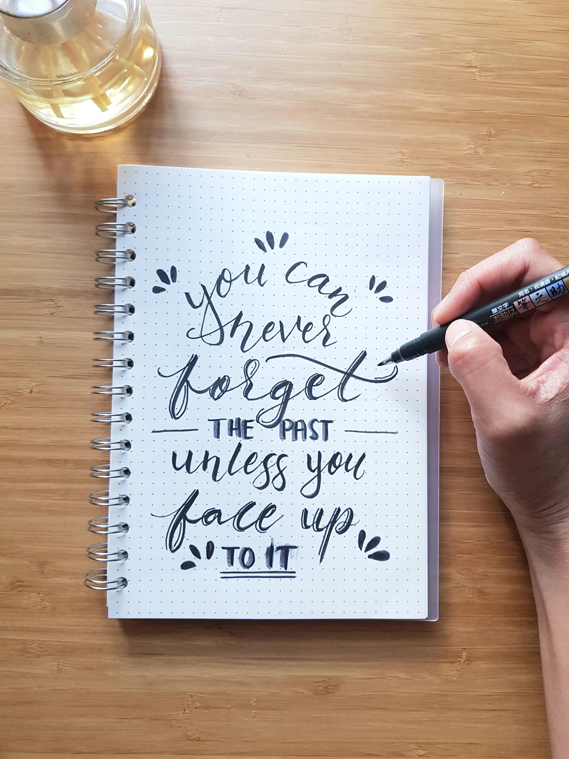 Handwriting in a spiral notebook on a wooden table, with a hand holding a pen and a drink nearby.