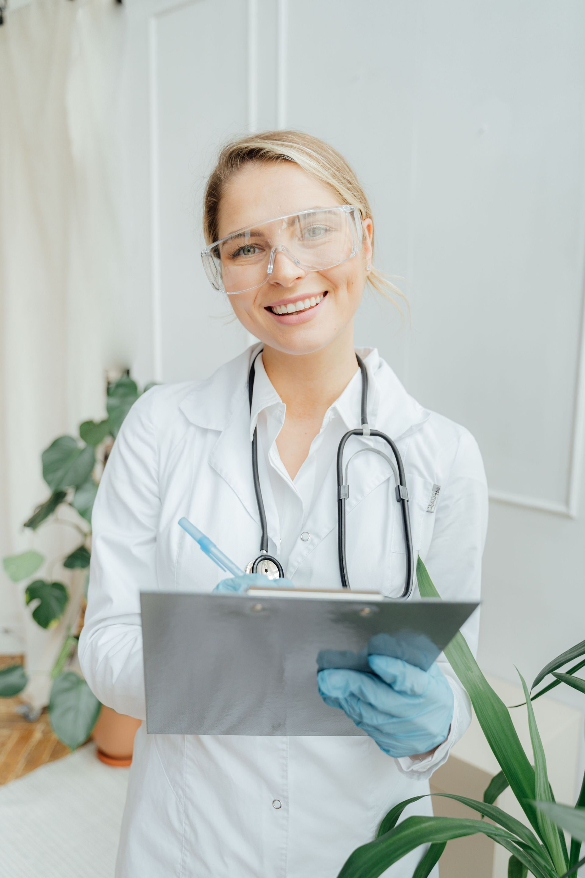 Smiling doctor in white coat and gloves holding a clipboard with a stethoscope around the neck