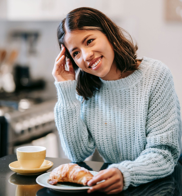 Smiling person in a light blue sweater at a table with coffee and a croissant in a bright kitchen