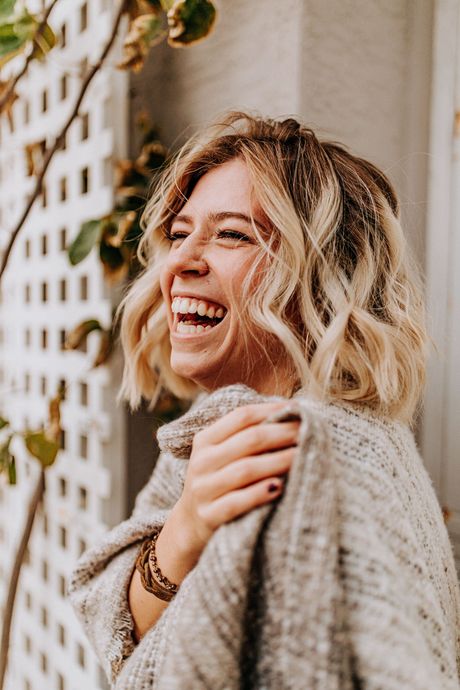Smiling woman with wavy blonde hair in a cozy sweater, standing outdoors by a lattice wall