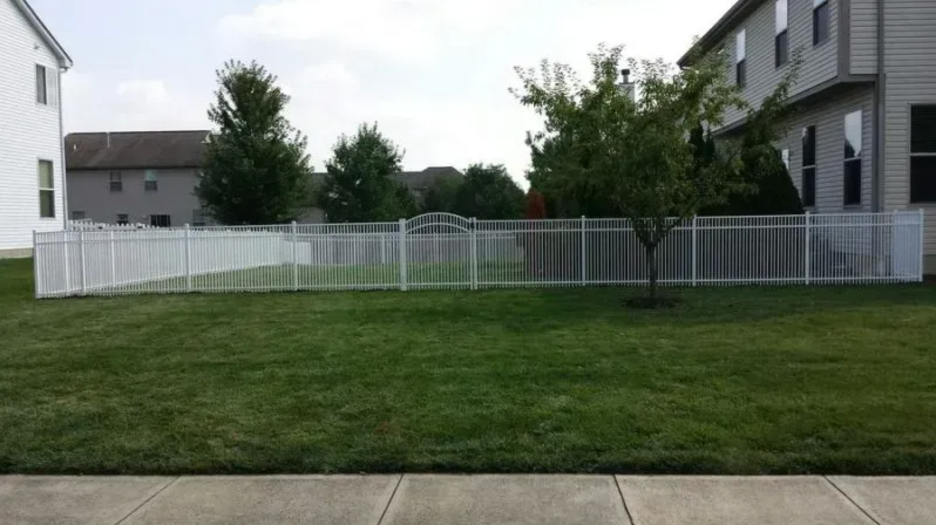 A white aluminum fence encloses a green grassy backyard between two residential houses on a sunny day in Columbus