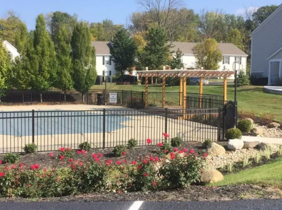 A fenced-in swimming pool with a covered wooden pergola, surrounded by red rose bushes and trees on a sunny day in Columbus