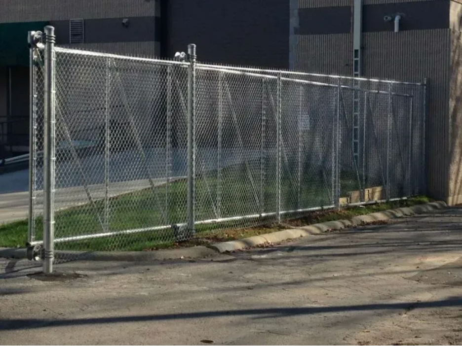 A silver chain-link fence stands in front of a commercial building, bordering a patch of grass and an asphalt lot in Columbus