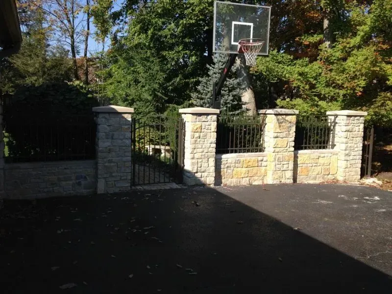 A basketball hoop is in the middle of a driveway surrounded by stone pillars and aluminum fence in Columbus