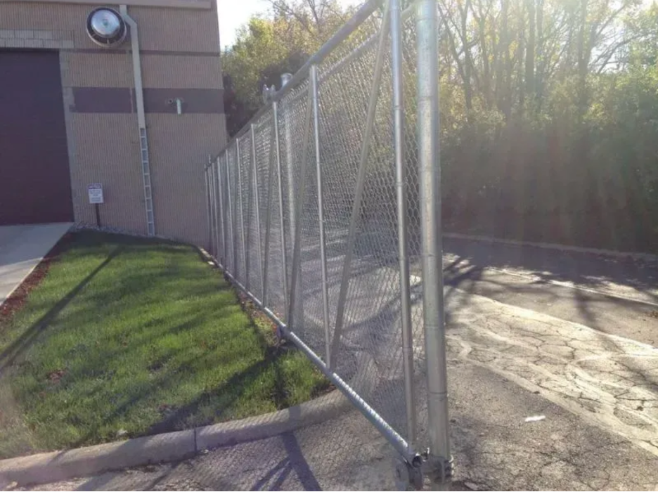 A chain-link fence extends from the side of a tan building toward a tree-lined asphalt area in Columbus