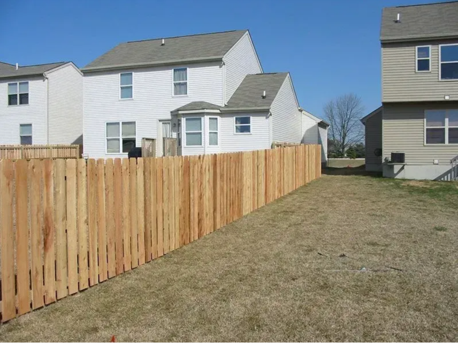 A long, wooden fence separates a grassy backyard from several suburban houses under a clear blue sky in Columbus