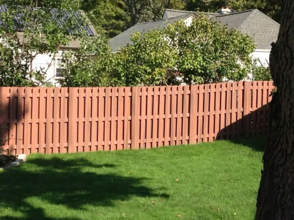 A reddish-brown wooden privacy fence stands in a grassy backyard, with a tree trunk in the foreground and houses behind in Columbus