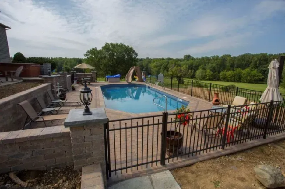 A blue rectangular swimming pool with a slide, surrounded by a stone patio, lounge chairs, and a metal fence in Columbus