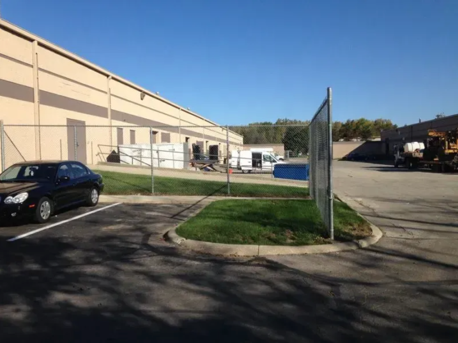 A black car parked in a paved lot next to a tan commercial building, with a chain-link fence and green grass in view in Columbus