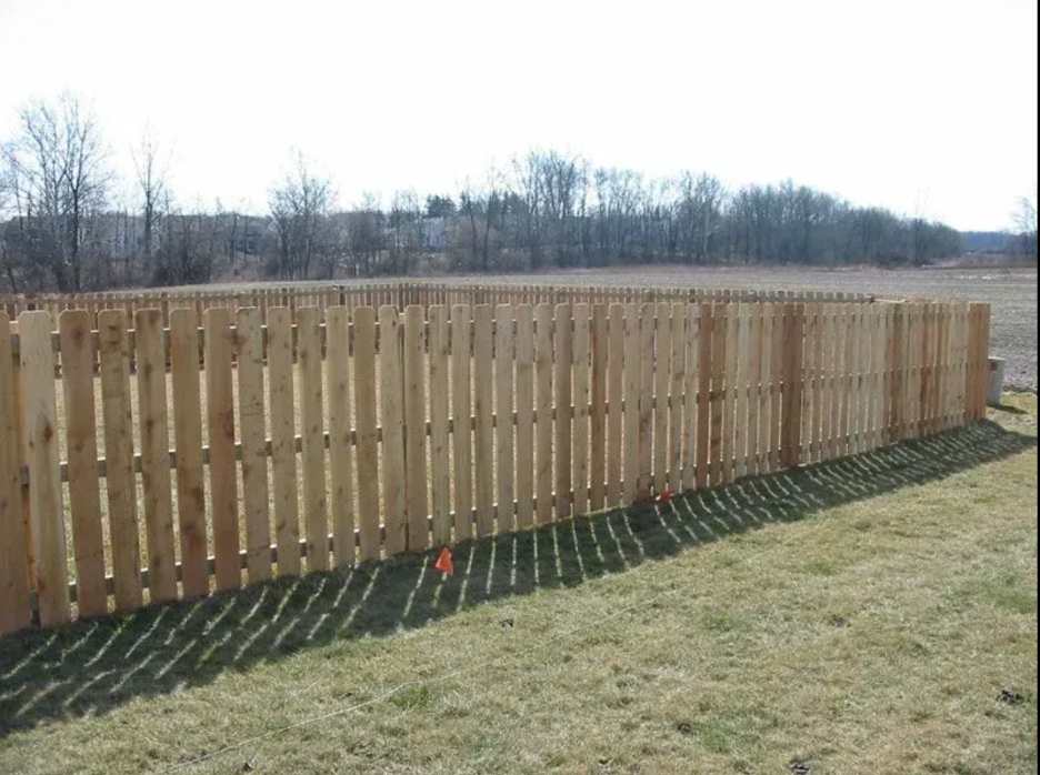 A new wooden picket fence stands in a grassy field on a bright day, with small orange flags marking the ground in Columbus