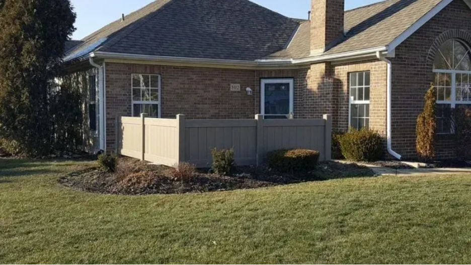 A suburban brick house exterior with a tan privacy fence surrounding a small landscaped garden bed on the front lawn in Columbus