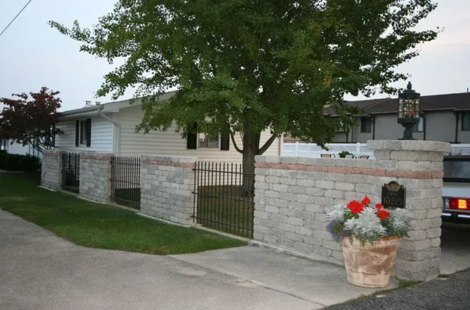 A house with light-colored siding behind a stone and wrought-iron fence, featuring a large tree and a potted flower urn in Columbus