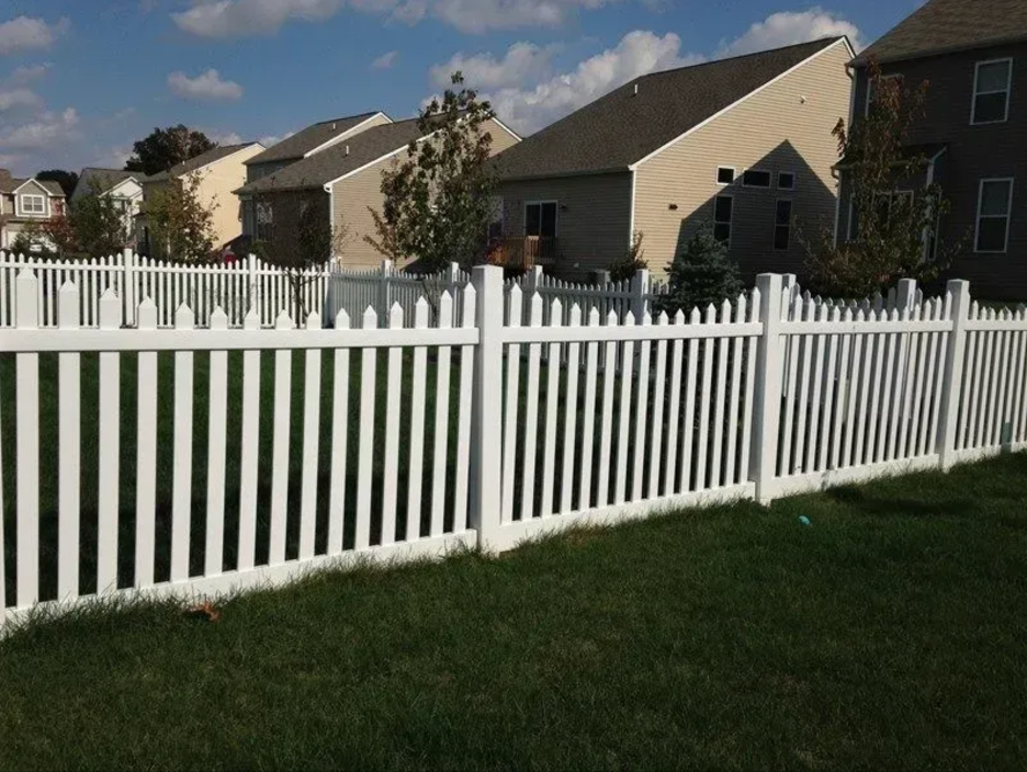 A white vinyl picket fence stands in a grassy yard in front of several suburban homes under a blue sky with clouds in Columbus