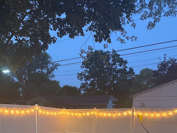 A string of warm, glowing patio lights draped along a white vinyl fence against a twilight sky with trees in the background in Columbus