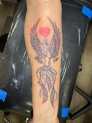 A person has a tattoo of a phoenix with a heart on their forearm.