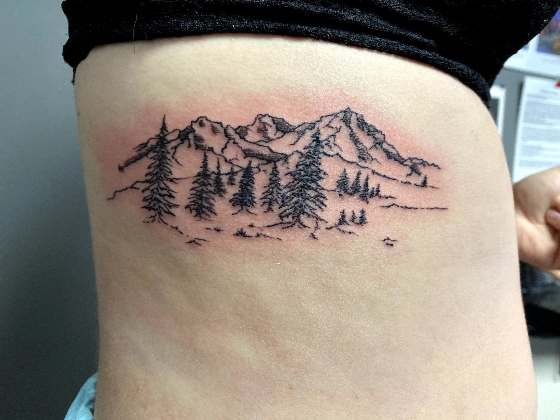 A woman has a tattoo of mountains and trees on her side.