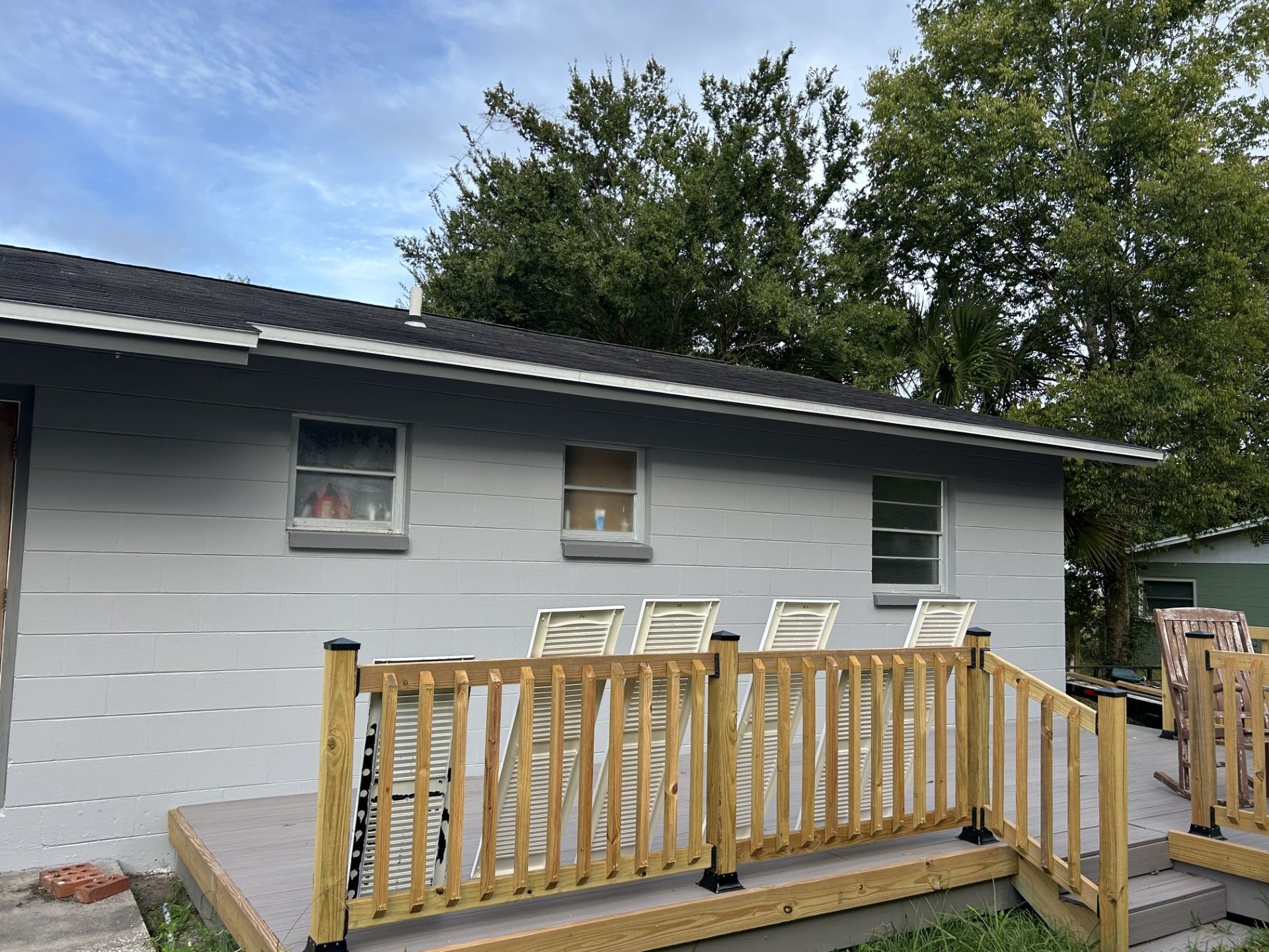 A grey house exterior with three windows and a wooden deck featuring a railing and bar-style chairs.