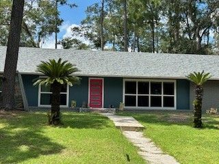 A single-story, blue-gray home with a bright pink door, a gray roof, and two small palm trees on a sunny grassy lawn.