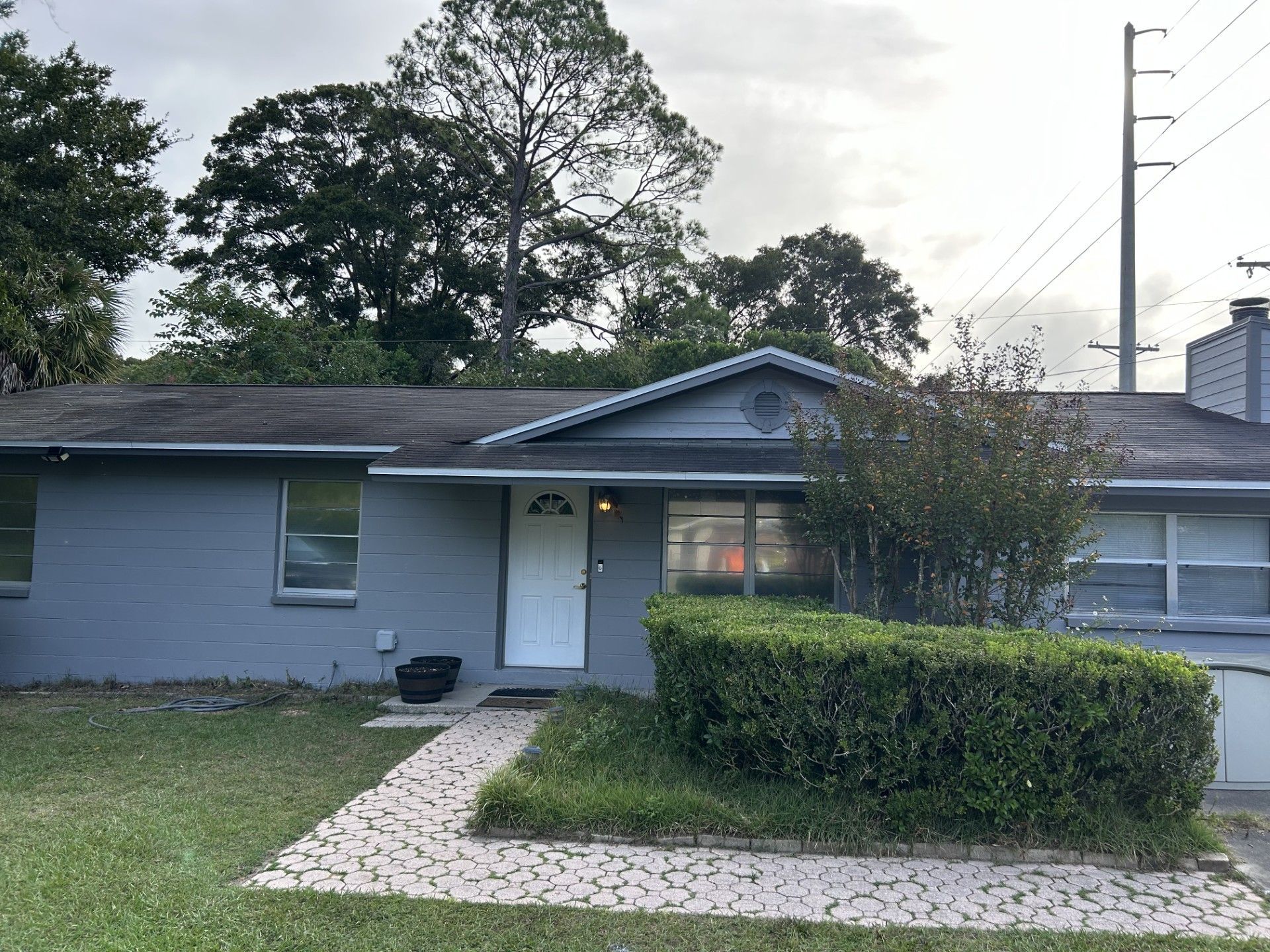 A single-story gray house with a white door, a paved stone walkway, and a large green hedge in the front yard.