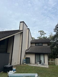 A light-colored two-story house with dark roof shingles and dark trim, featuring a prominent chimney and a rear patio.