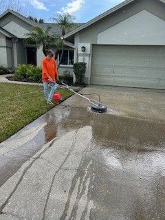 A person in an orange shirt uses a circular pressure washer to clean a concrete driveway in front of a house.