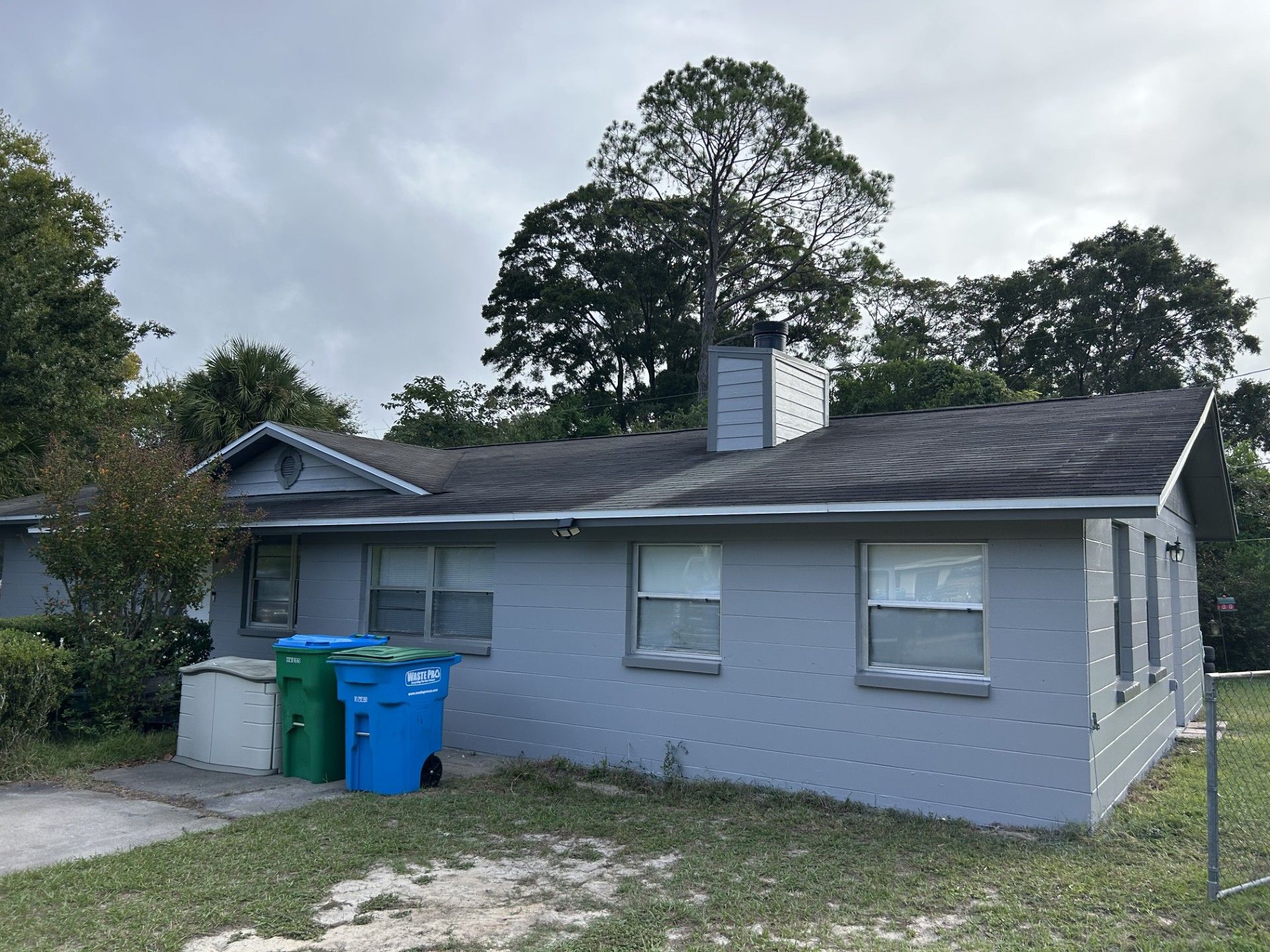 A single-story, light-gray house with a dark shingled roof, a chimney, and two trash cans parked near the side wall.