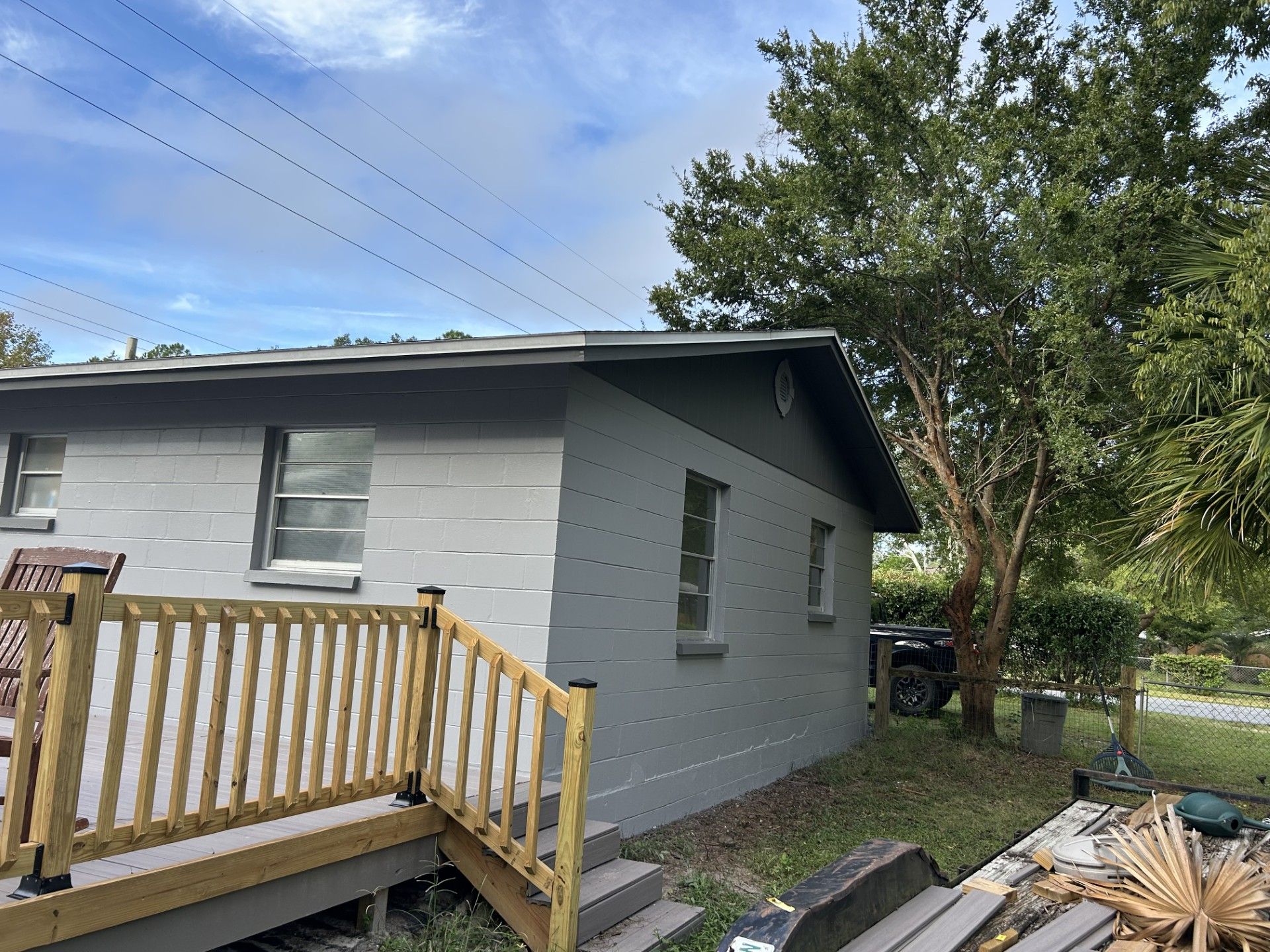 A grey-painted house with a wood deck and railing stands under a blue sky next to a large tree.
