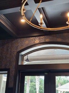 A gold circular chandelier hangs below a dark coffered ceiling, positioned above an arched doorway looking onto a patio.