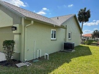 A light green, single-story house exterior under a blue sky, featuring a side window, AC unit, and a tall palm tree.