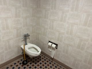 A wall-mounted toilet with a plunger next to it and a double toilet paper dispenser on the tiled bathroom wall.